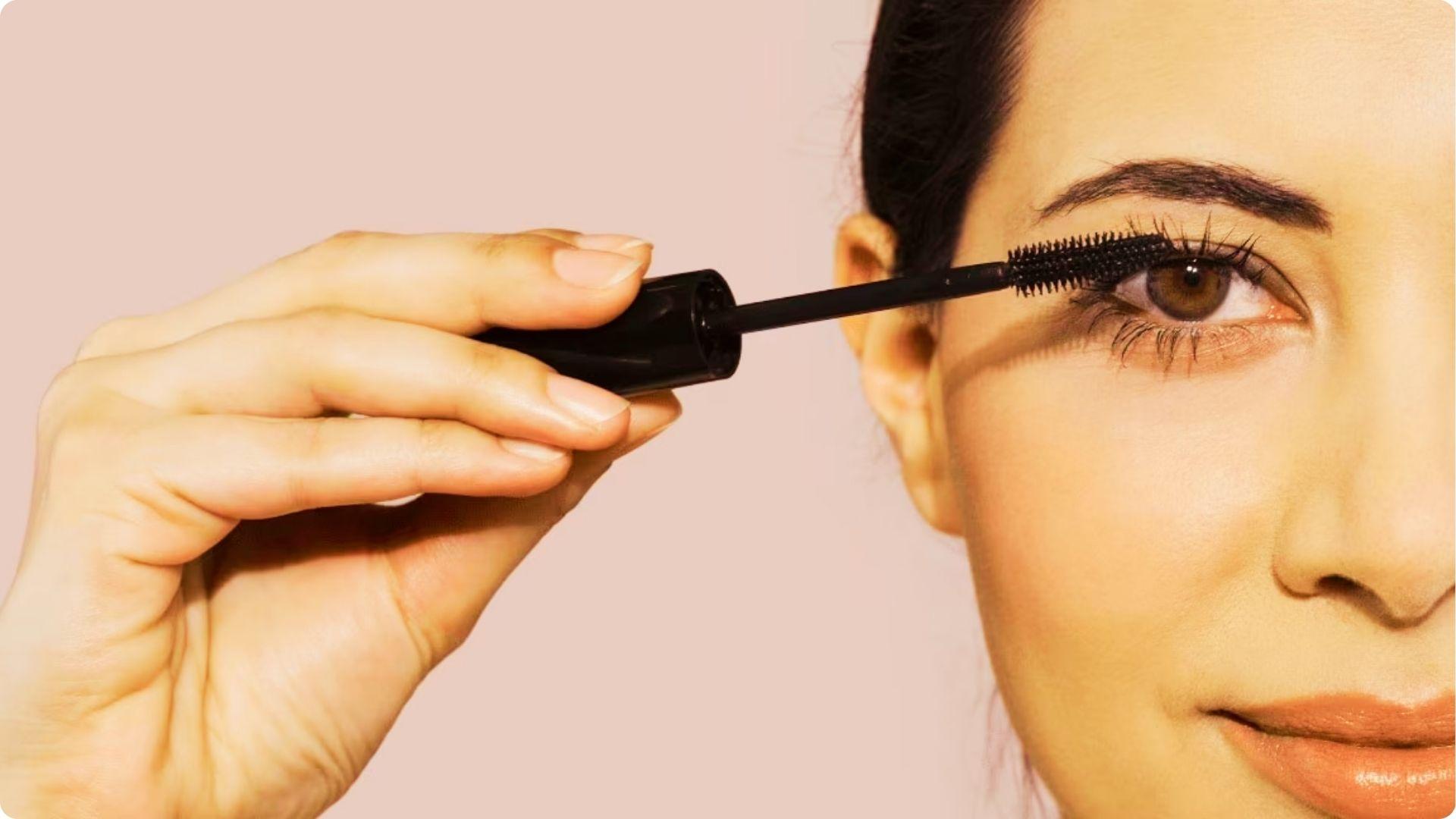 Hand holding a black mascara wand applying product to upper lashes against a soft pink background.