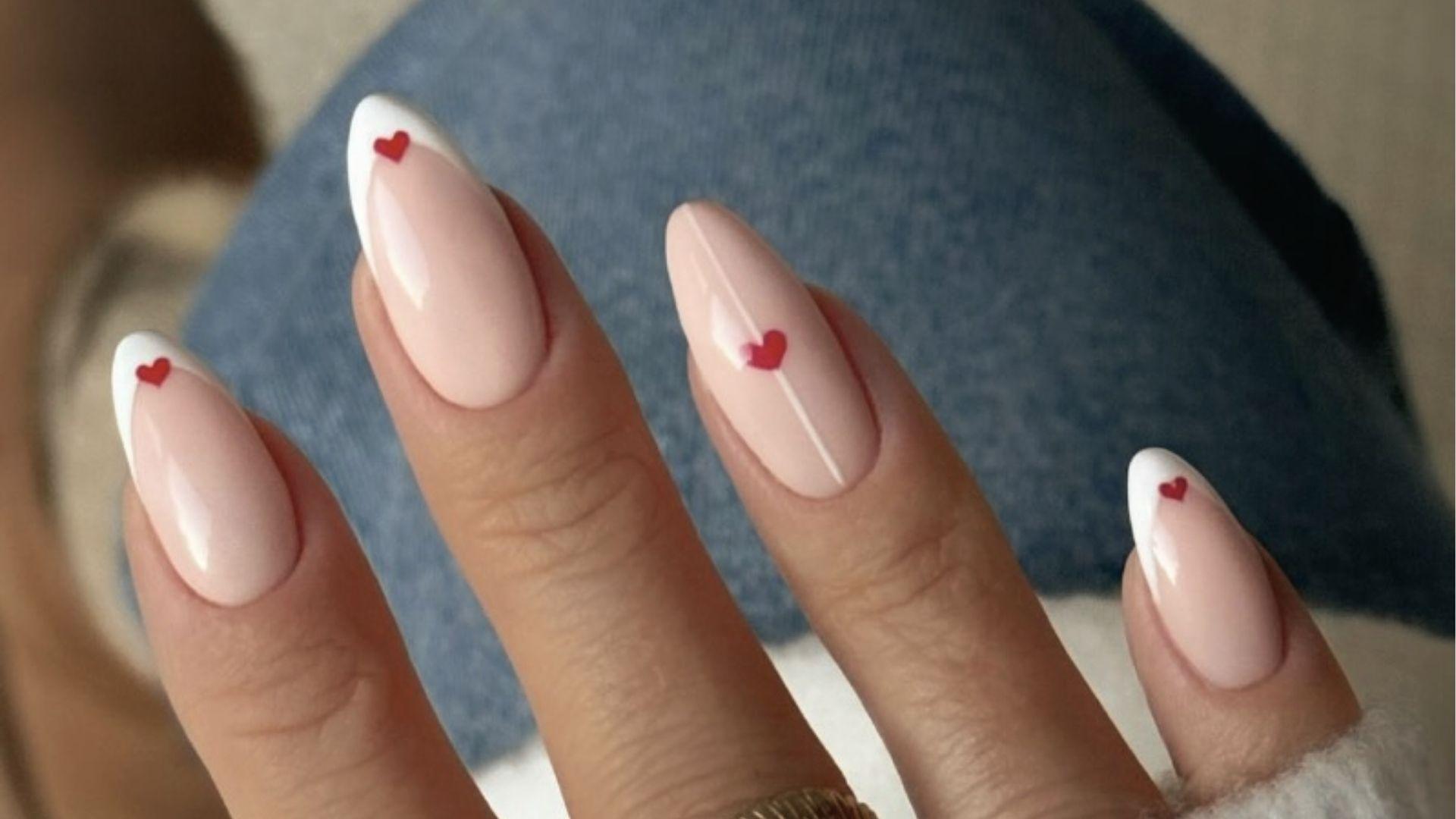Short almond dip nails with glossy nude base, white French tips and tiny red heart details on top.
