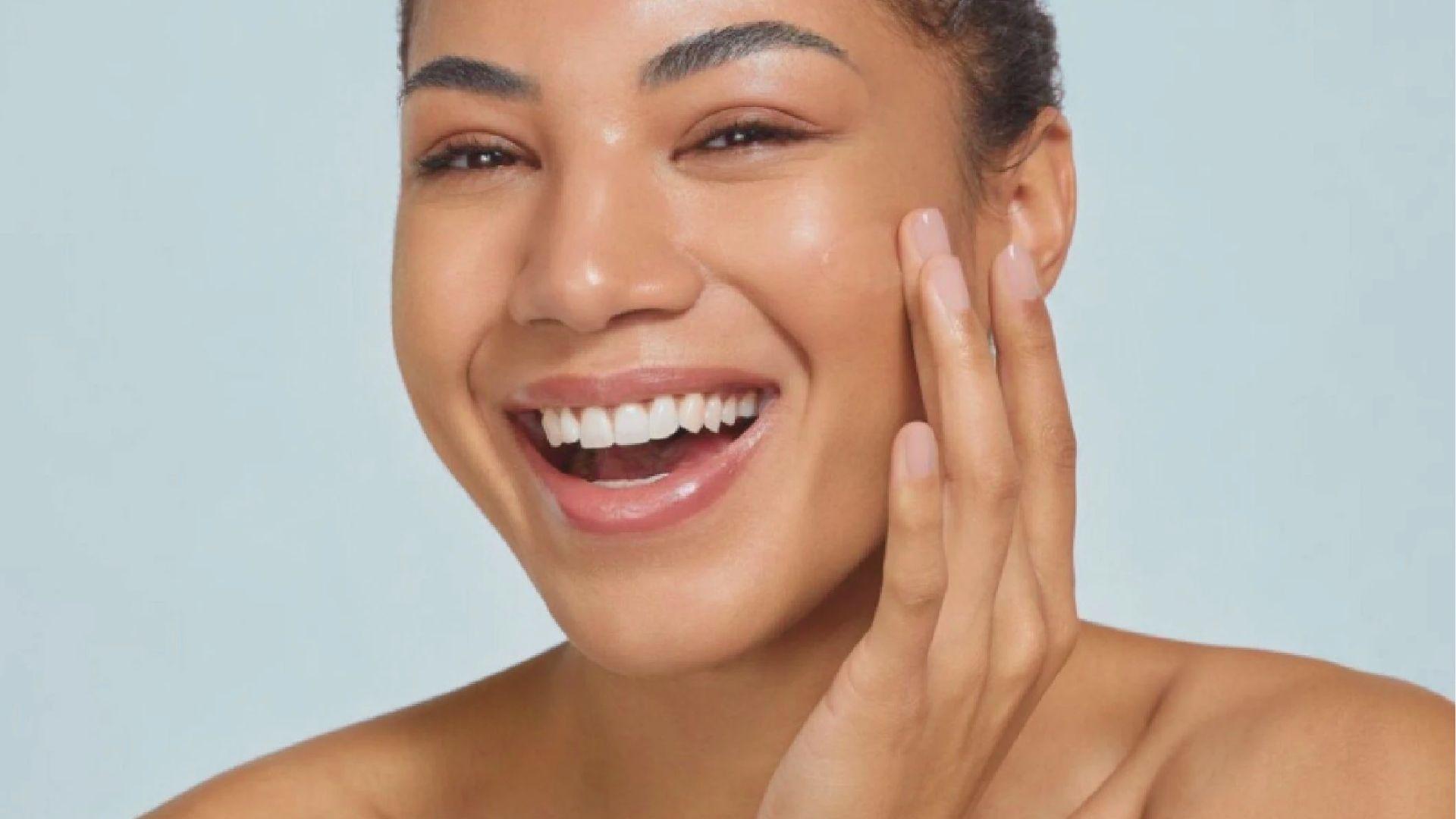 A joyful woman with a radiant smile touches her smooth, glowing cheek against a light blue background.