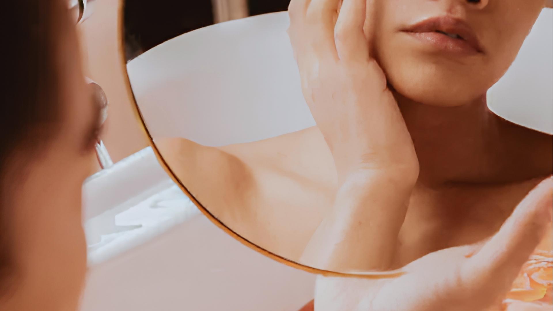 Woman looks in round mirror while touching jawline and neck, examining tight skin texture caused by dehydration or over-exfoliation.