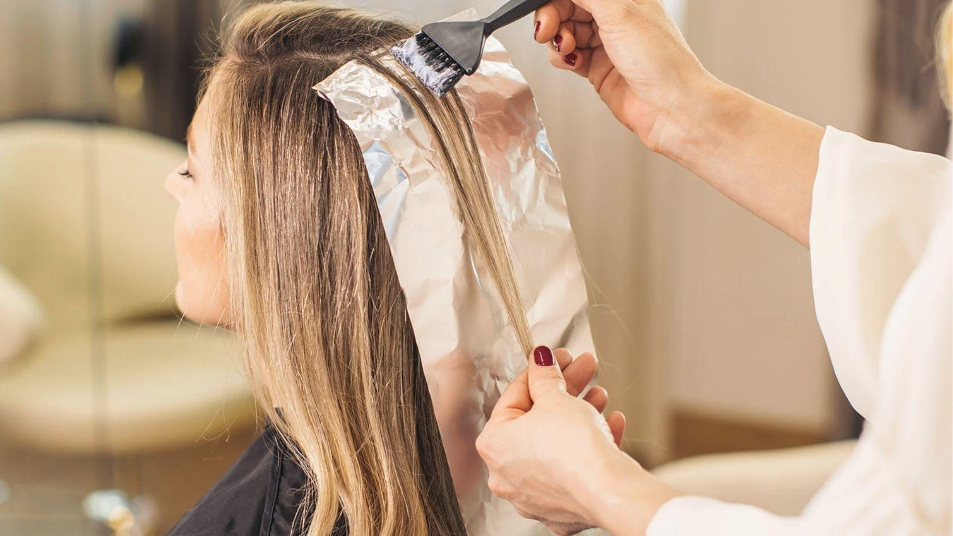 Brush applying bleach to dark roots with hair tied up, showing timed root touch-up application.