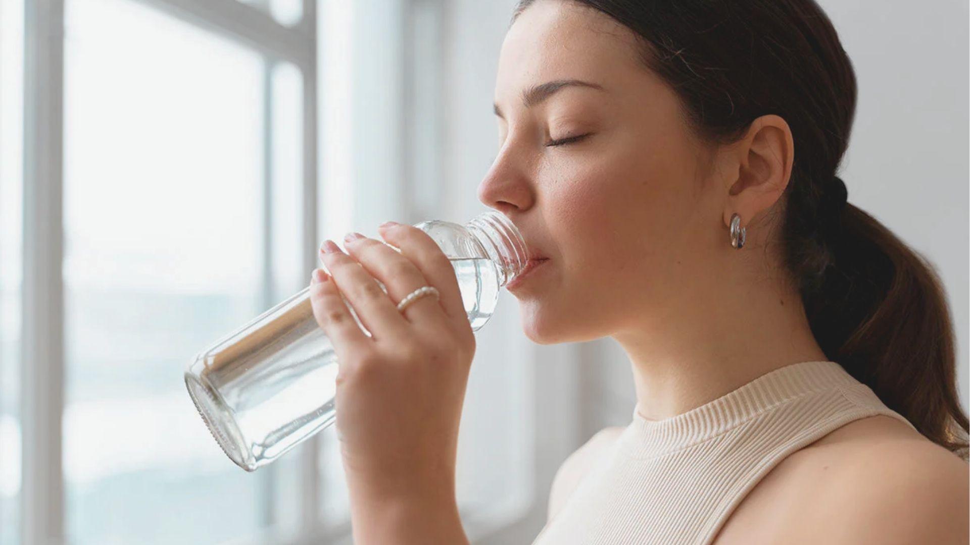 Side profile of a woman with a ponytail drinking water from a clear bottle near a window