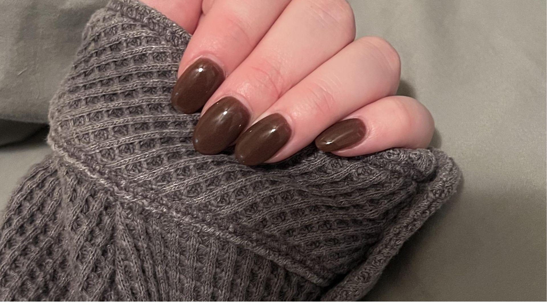 Short oval dip nails in glossy rich chocolate brown holding grey knit sweater for a cozy fall look.