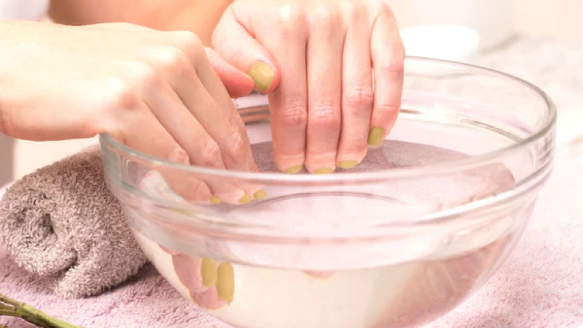 Hands with green polish soaking in clear glass bowl to remove gel nails using acetone for 15 minutes