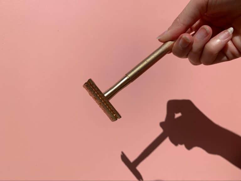 A hand holding a gold safety razor against a pink background. The shadow of the razor is cast clearly, creating a minimalist and clean aesthetic.