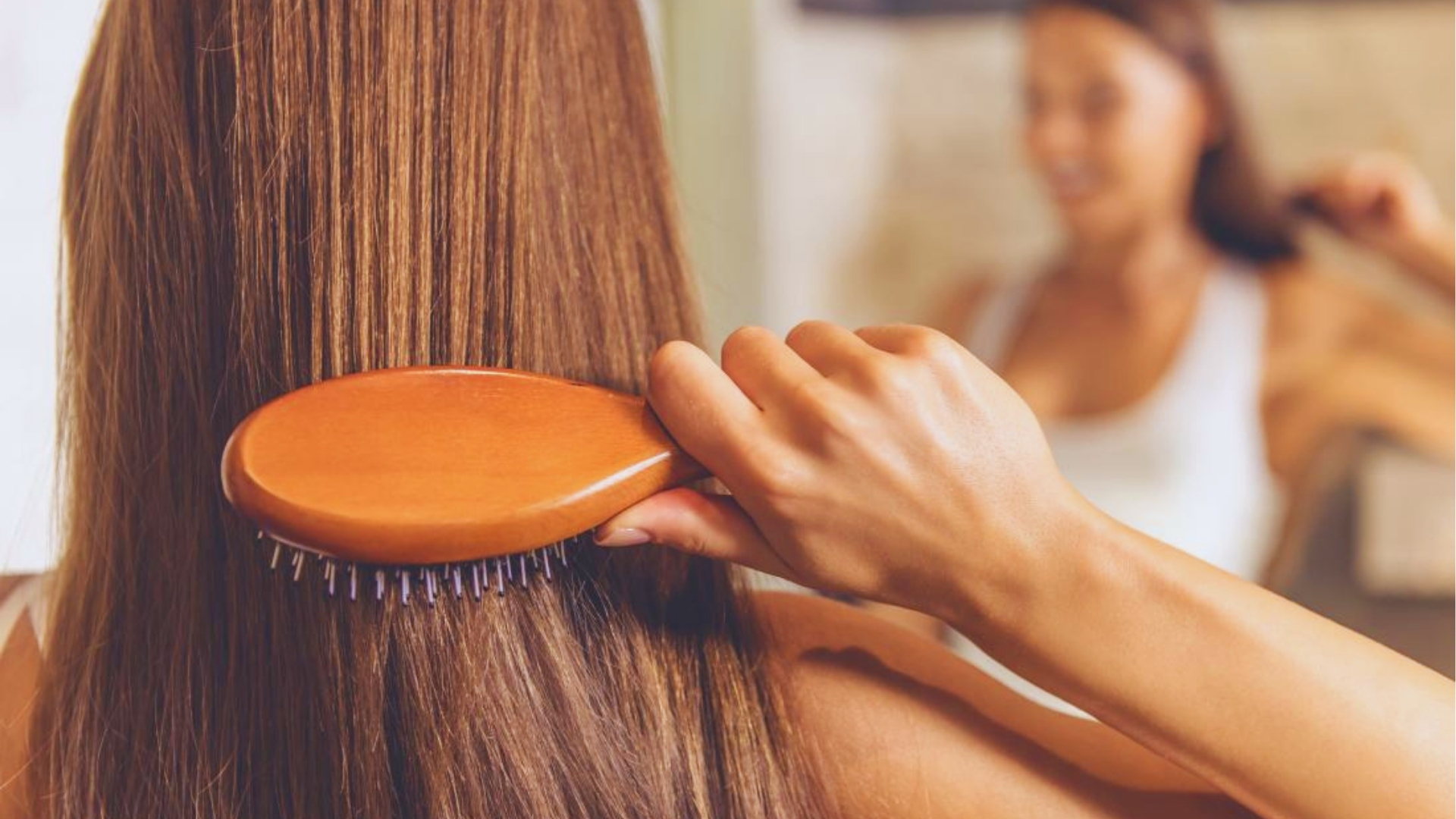 Back view of woman brushing long, straight, shiny brown hair with wooden brush, showing healthy results from oiling.