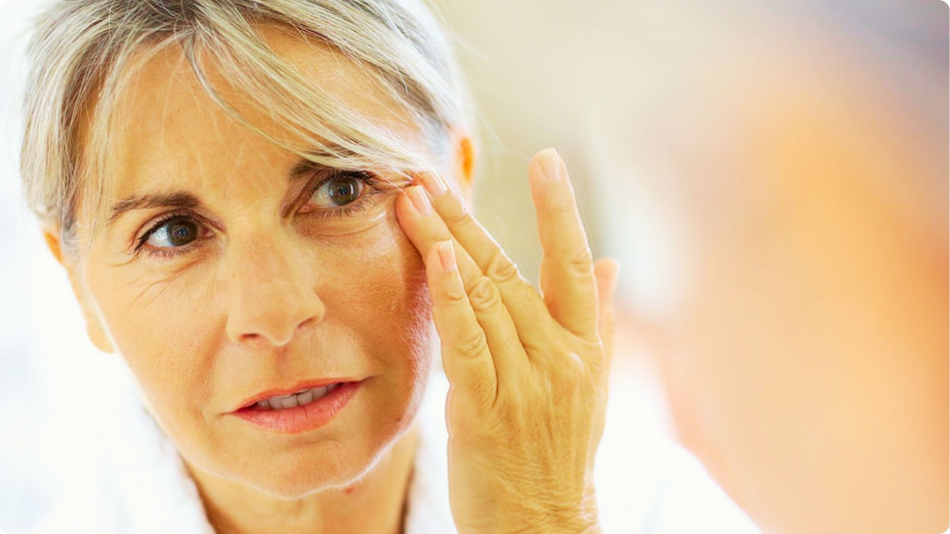 An older woman in a white robe gently touches fine lines around her eye in front of a mirror.