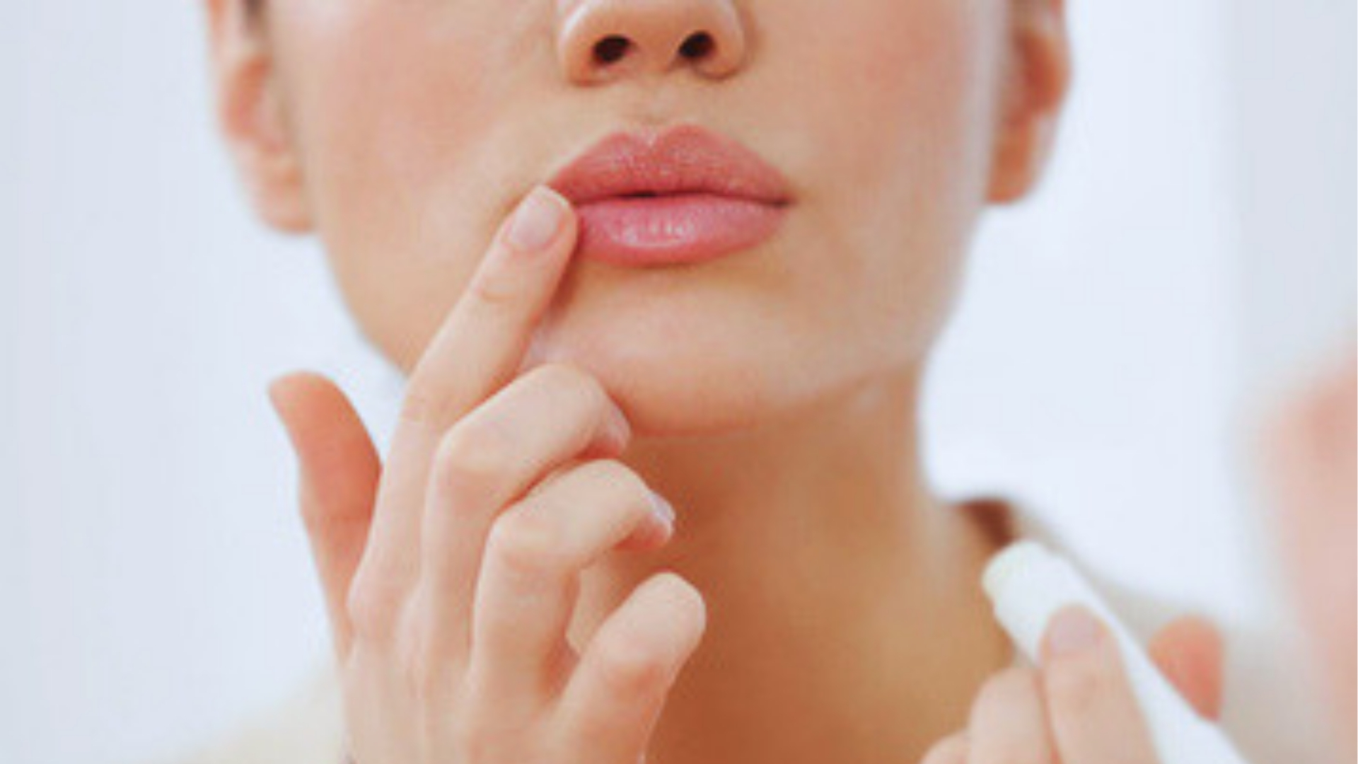 Woman applies clear lip balm from white tube to lips, showing lip plumper use for moisture and fullness.