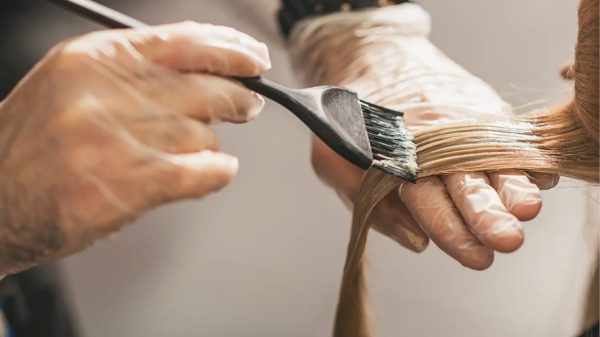 Gloved hands apply hair dye to light brown strands with brush. Semi permanent color lasts 4-6 weeks.