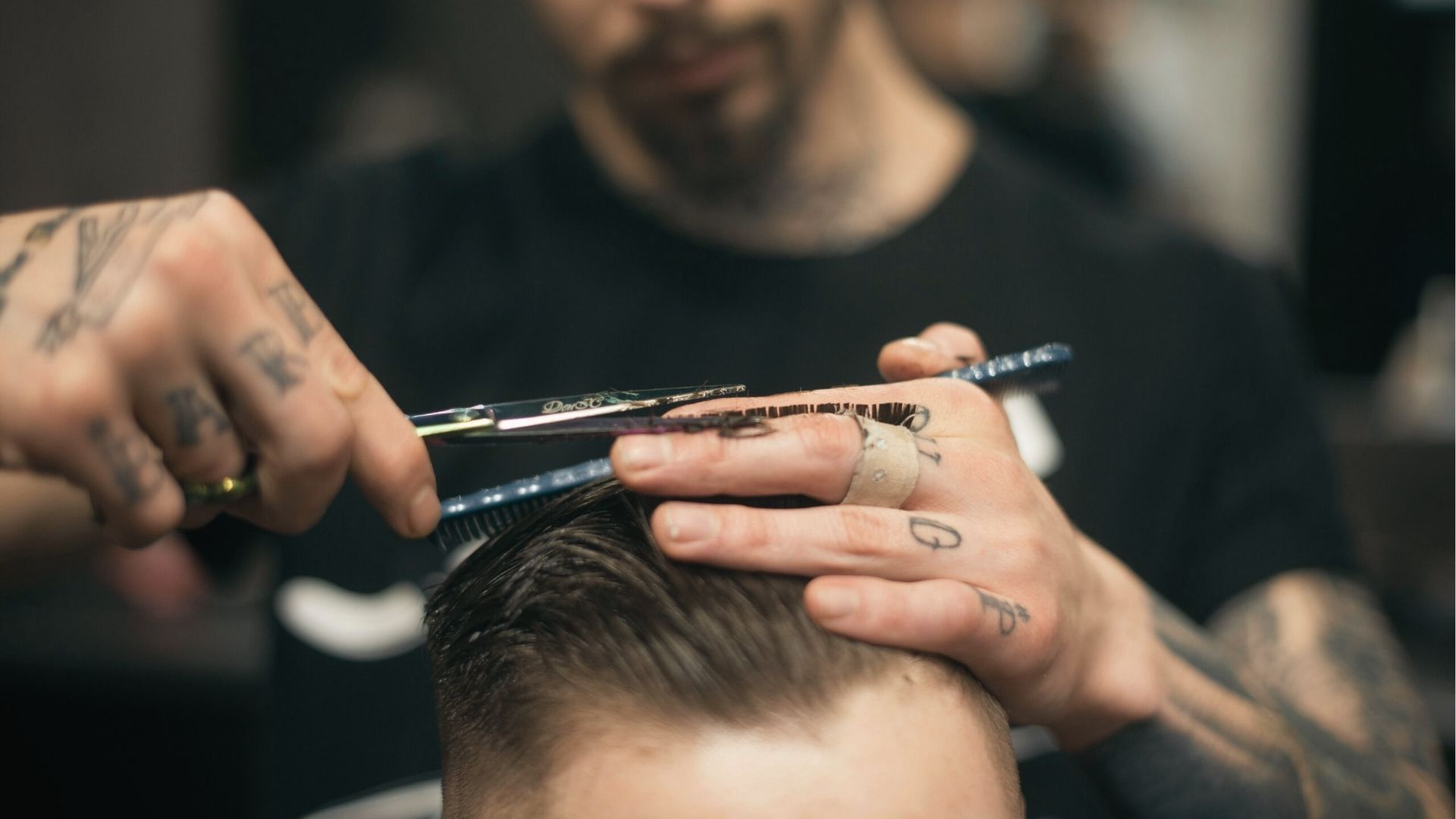 Tattooed barbers use scissors and comb to shape top hair while tapering sides for a clean fade cut.