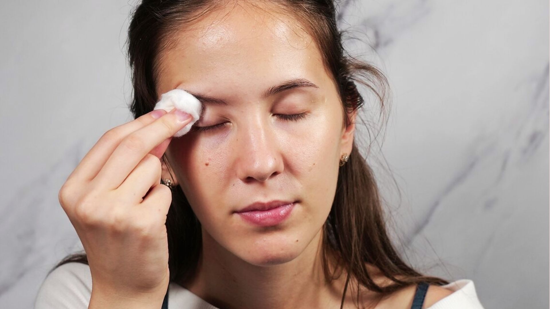Person gently cleaning the eyelid area with a cotton pad to prevent and reduce under-eye bumps.