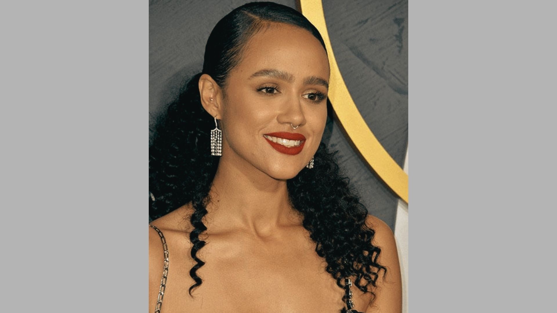 Smiling woman with long curly ponytail and red lipstick poses at event with gold backdrop