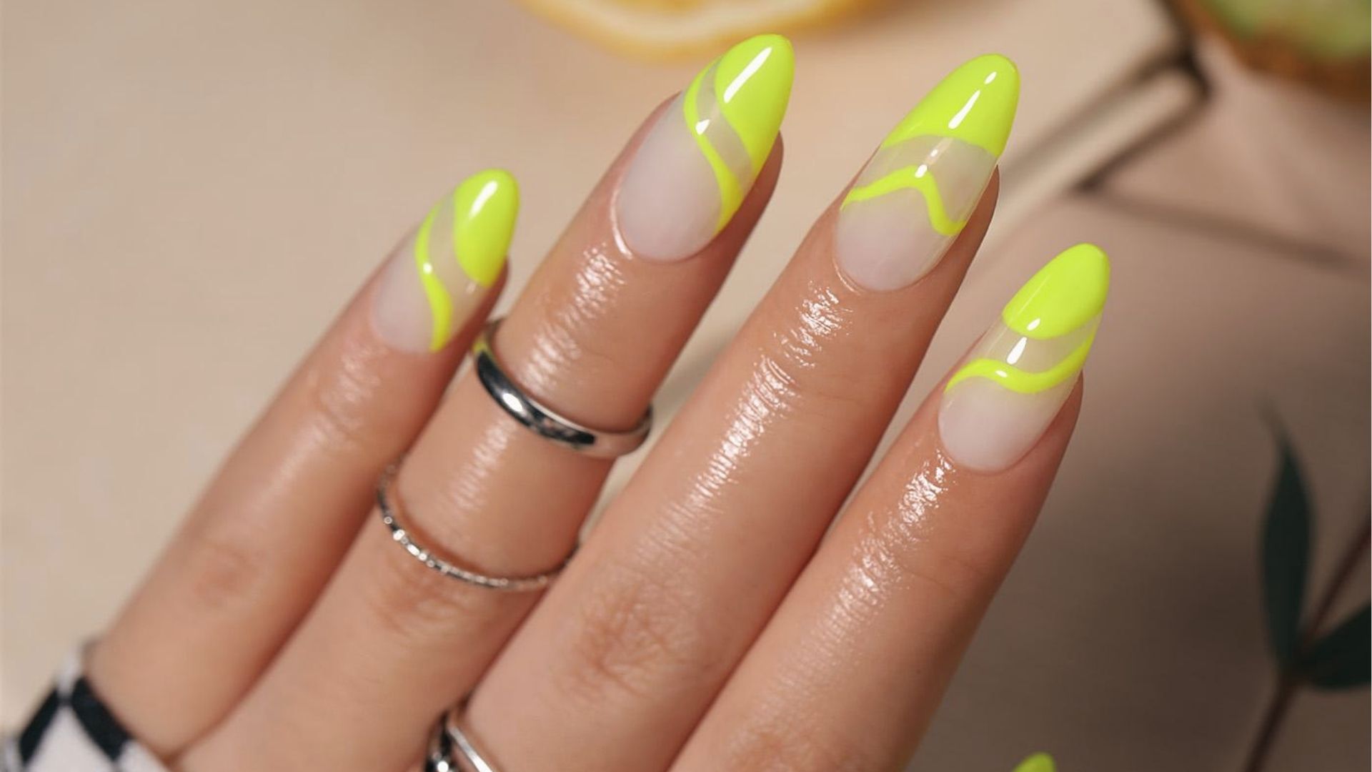 Almond nails with neon yellow swirls and negative space tips on nude base, glossy finish, silver rings