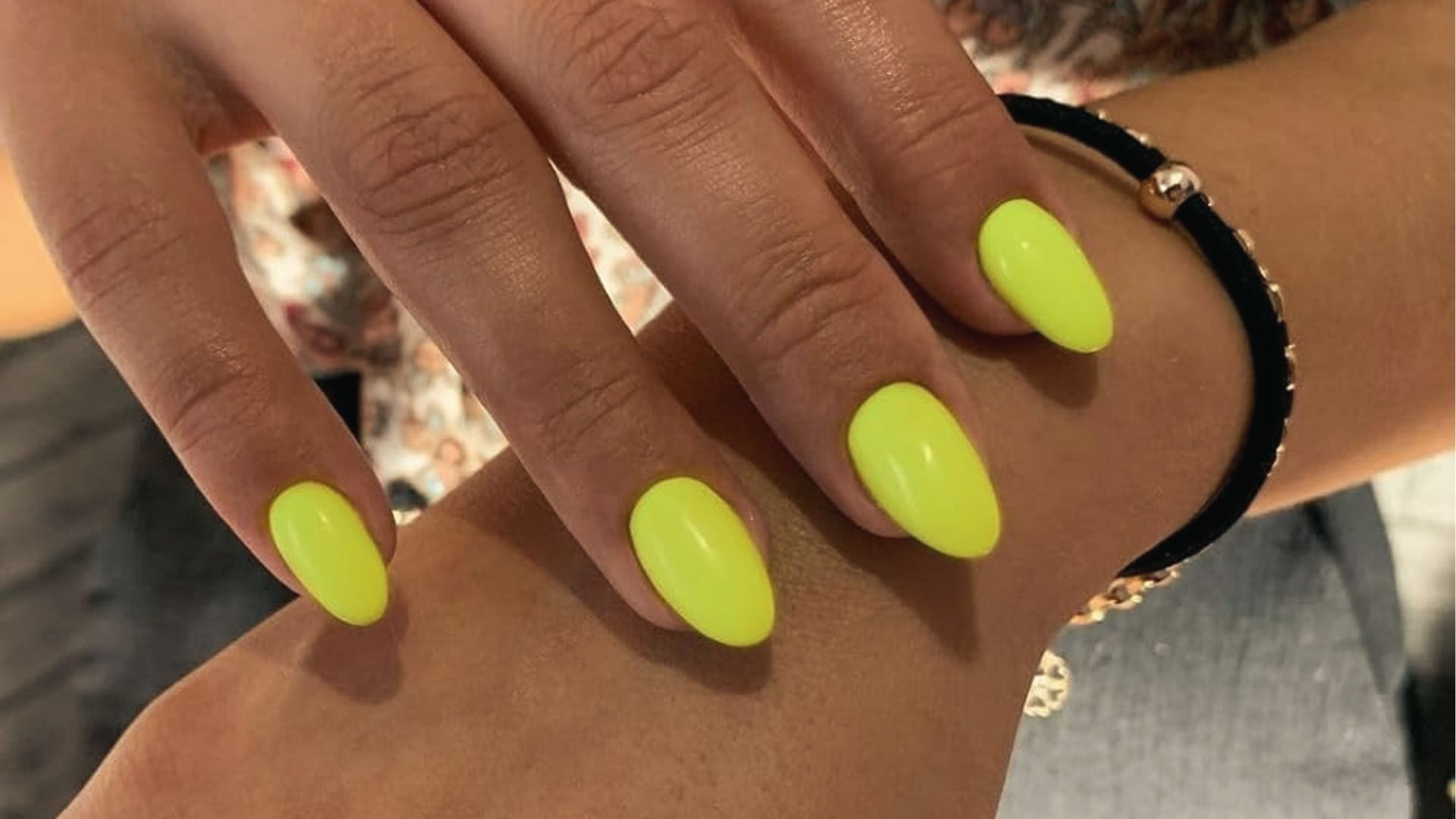 Oval nails in glossy neon yellow polish, hand with black hair tie bracelet and gold jewelry