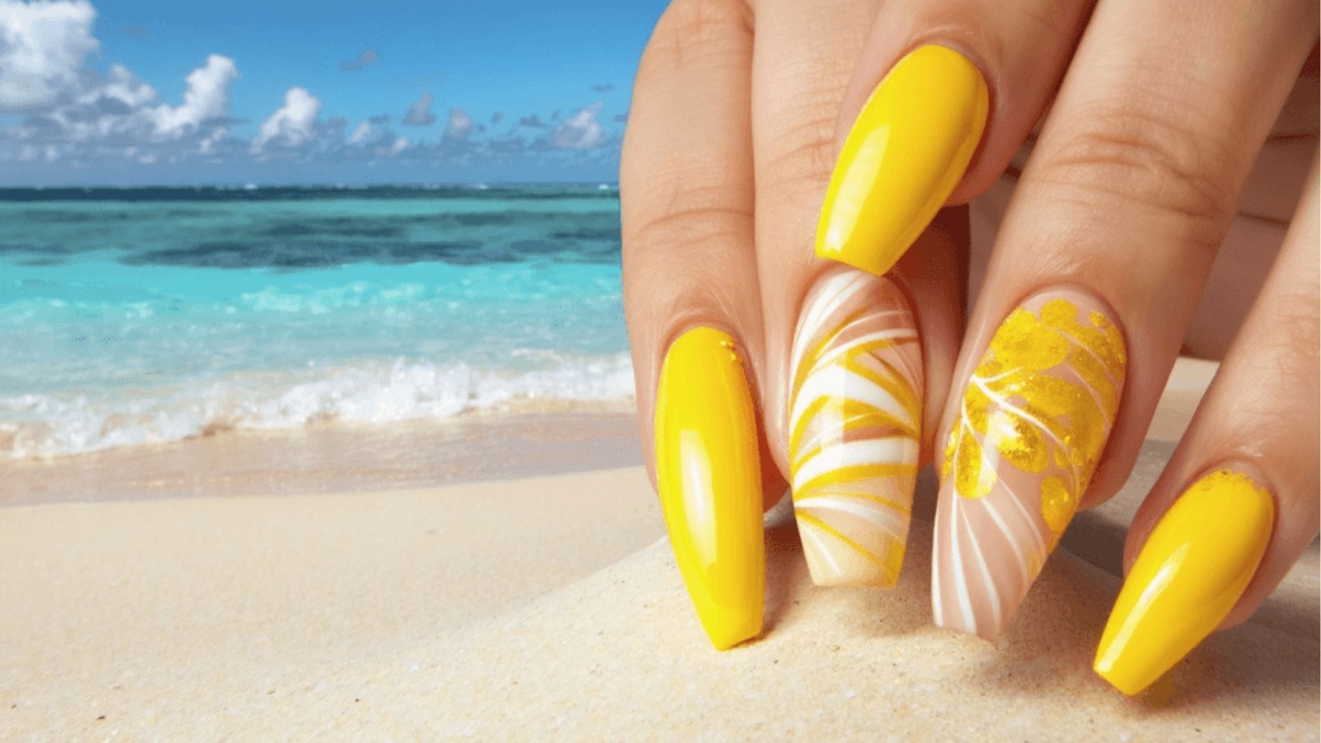 Long tropical nails in bright yellow neon with white swirls and gold leaf accents, posed on beach sand
