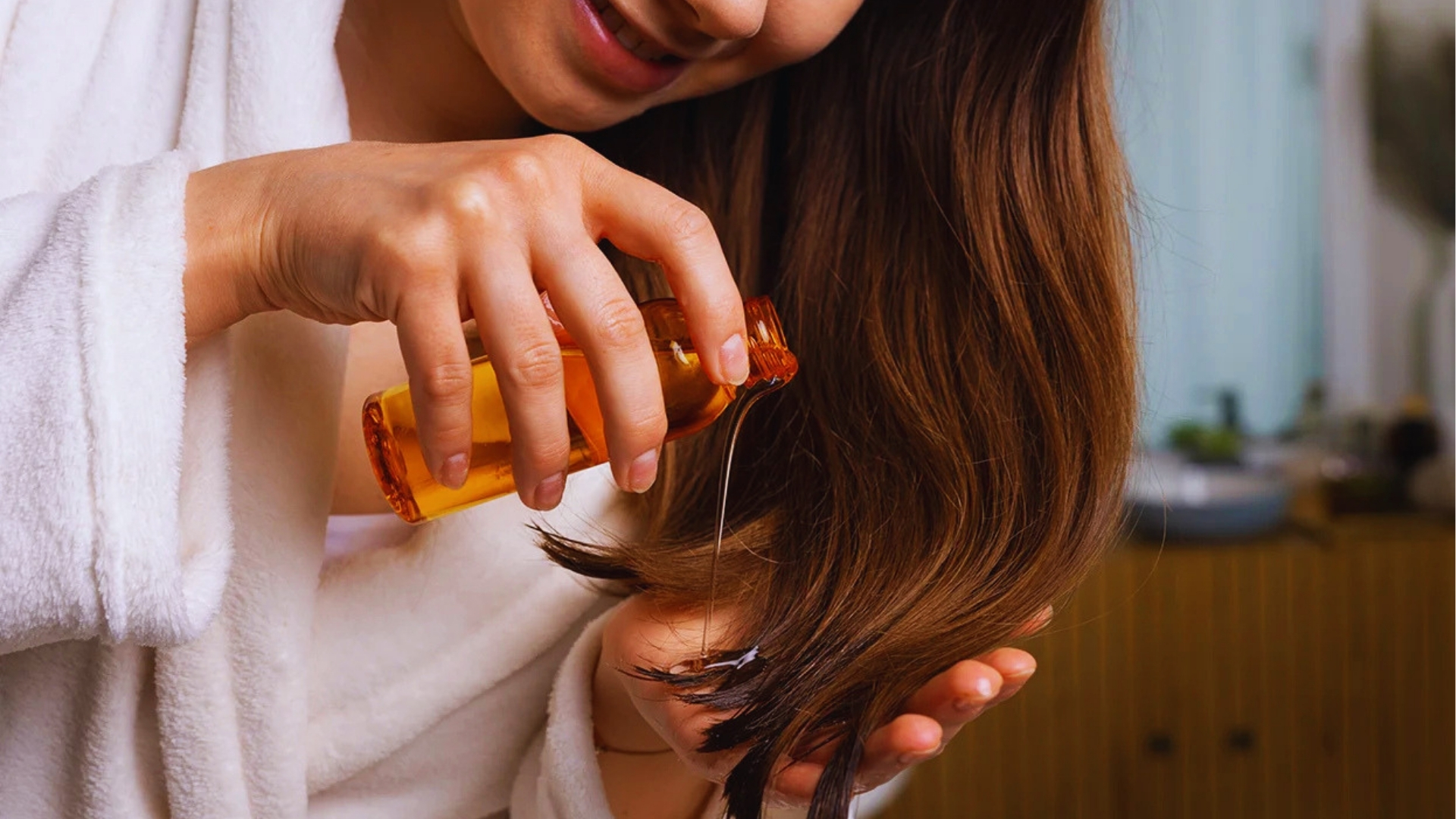 Woman in white robe pours amber vitamin E oil onto long brown hair ends to reduce dryness and split ends.
