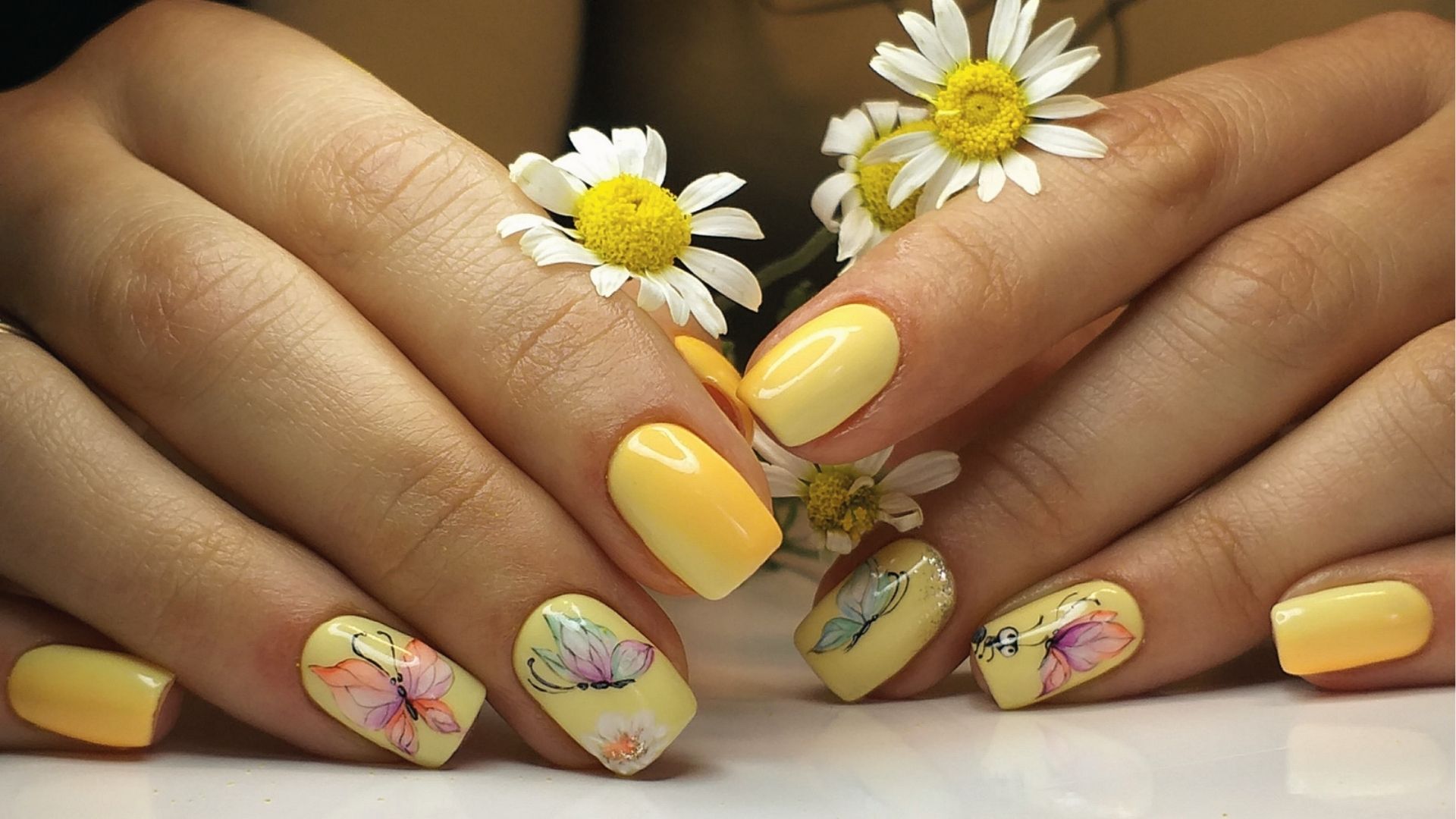 Short square yellow nails with watercolor butterfly art and daisies, hands holding white daisies