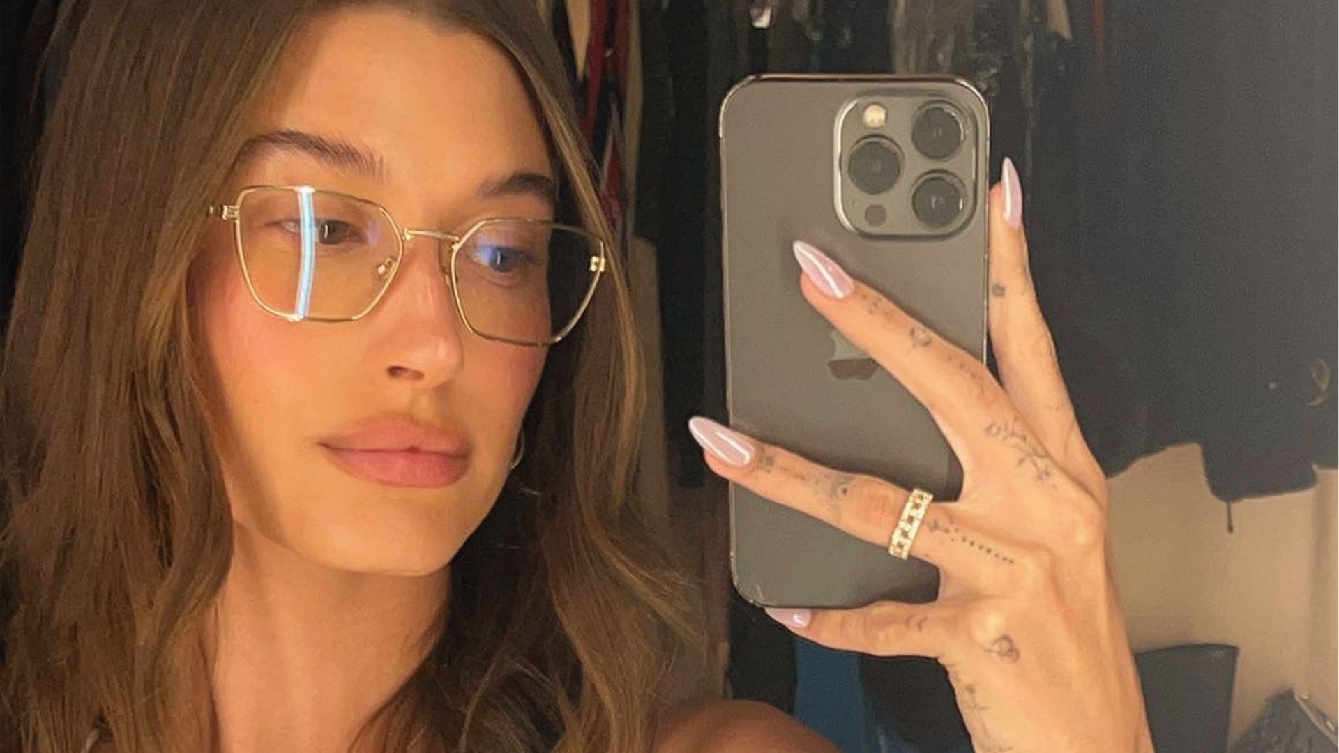 Hailey Bieber takes mirror selfie displaying long almond glazed donut nails with gold rings