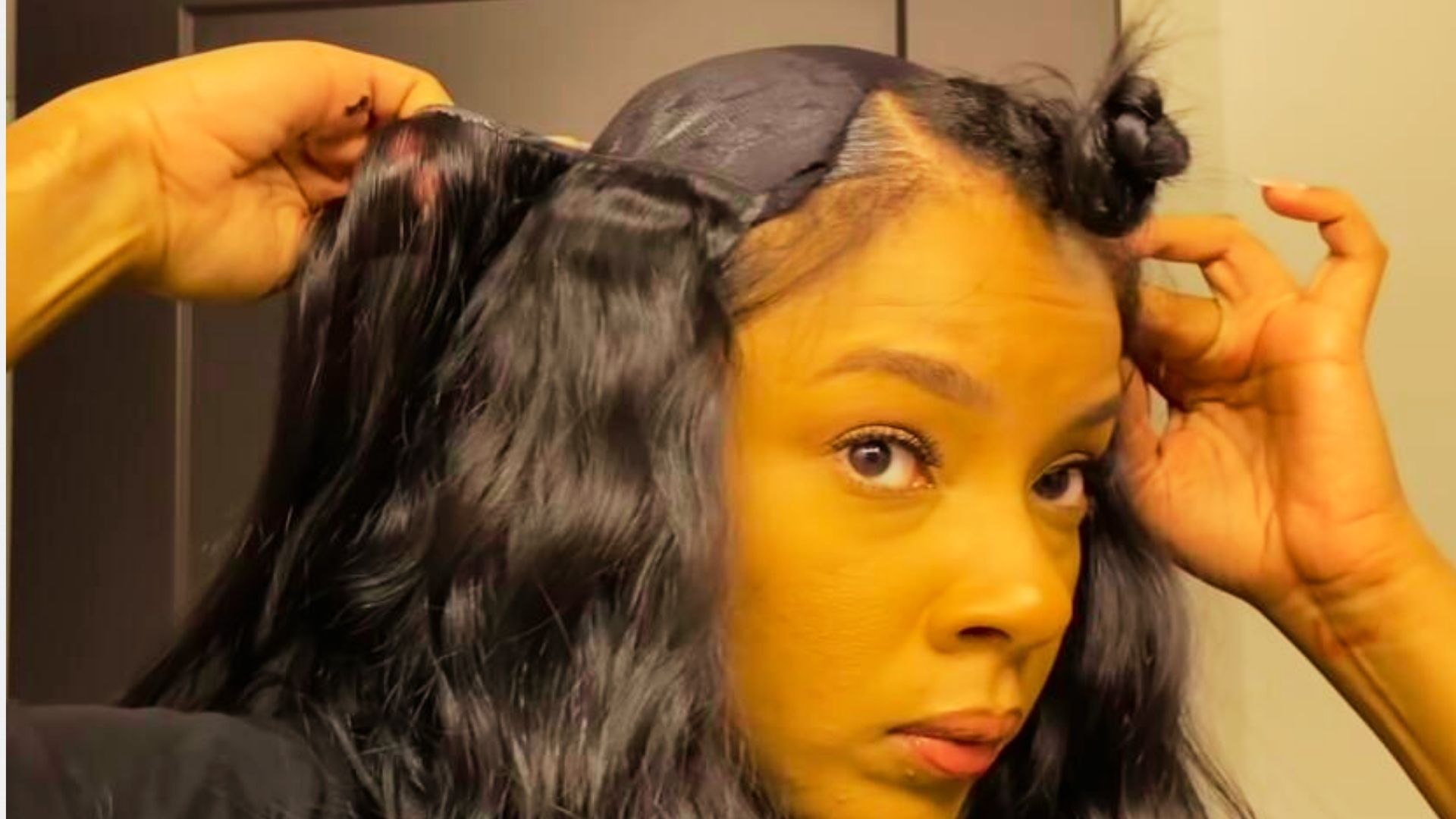 Woman installing wavy black hair extensions on wig cap while sectioning natural hair into bun
