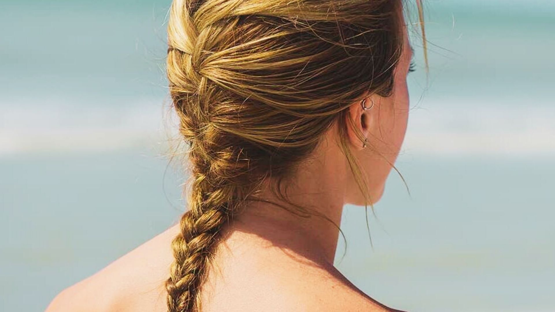Woman at beach with long loose French braid hairstyle and small earrings facing the ocean
