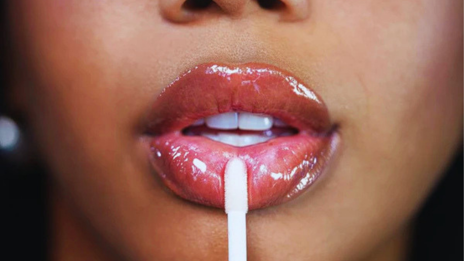 Close-up of glossy pink lips with applicator wand applying lip plumper gloss to create fuller lips instantly.