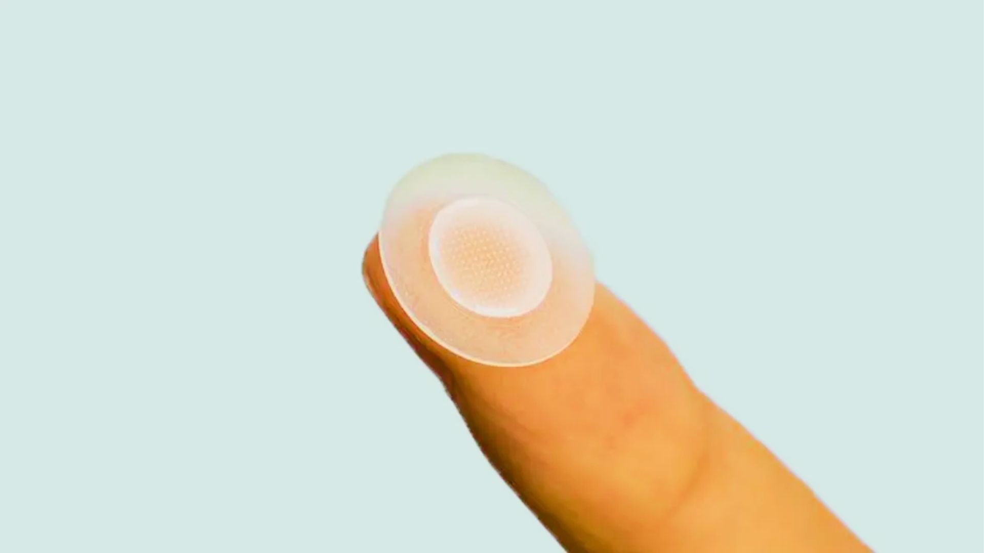 Closeup of fingertip holding round clear hydrocolloid pimple patch with microdots against light blue background