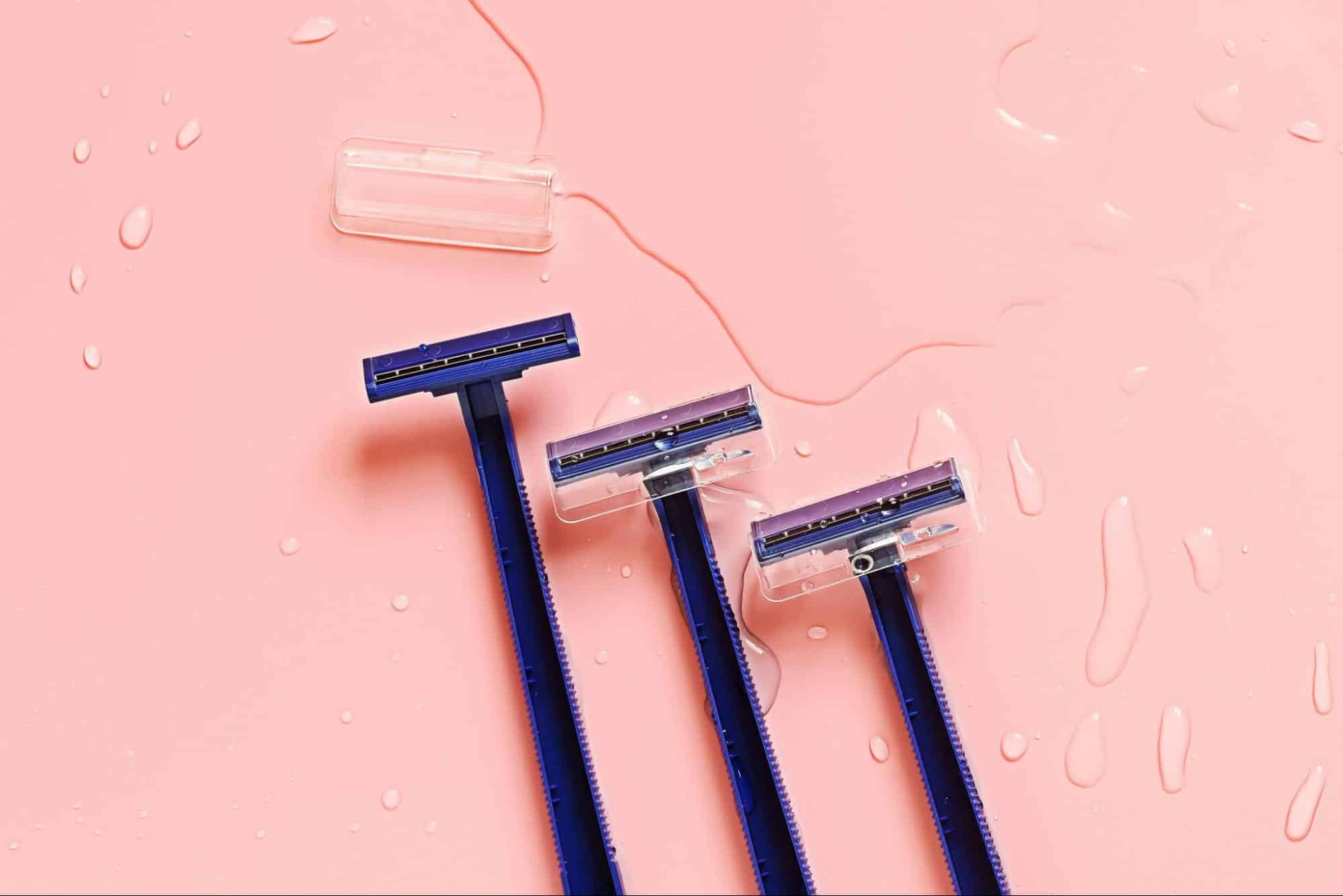Three dark blue razors with clear protective caps are arranged on a wet, pink surface, creating a fresh and clean aesthetic.