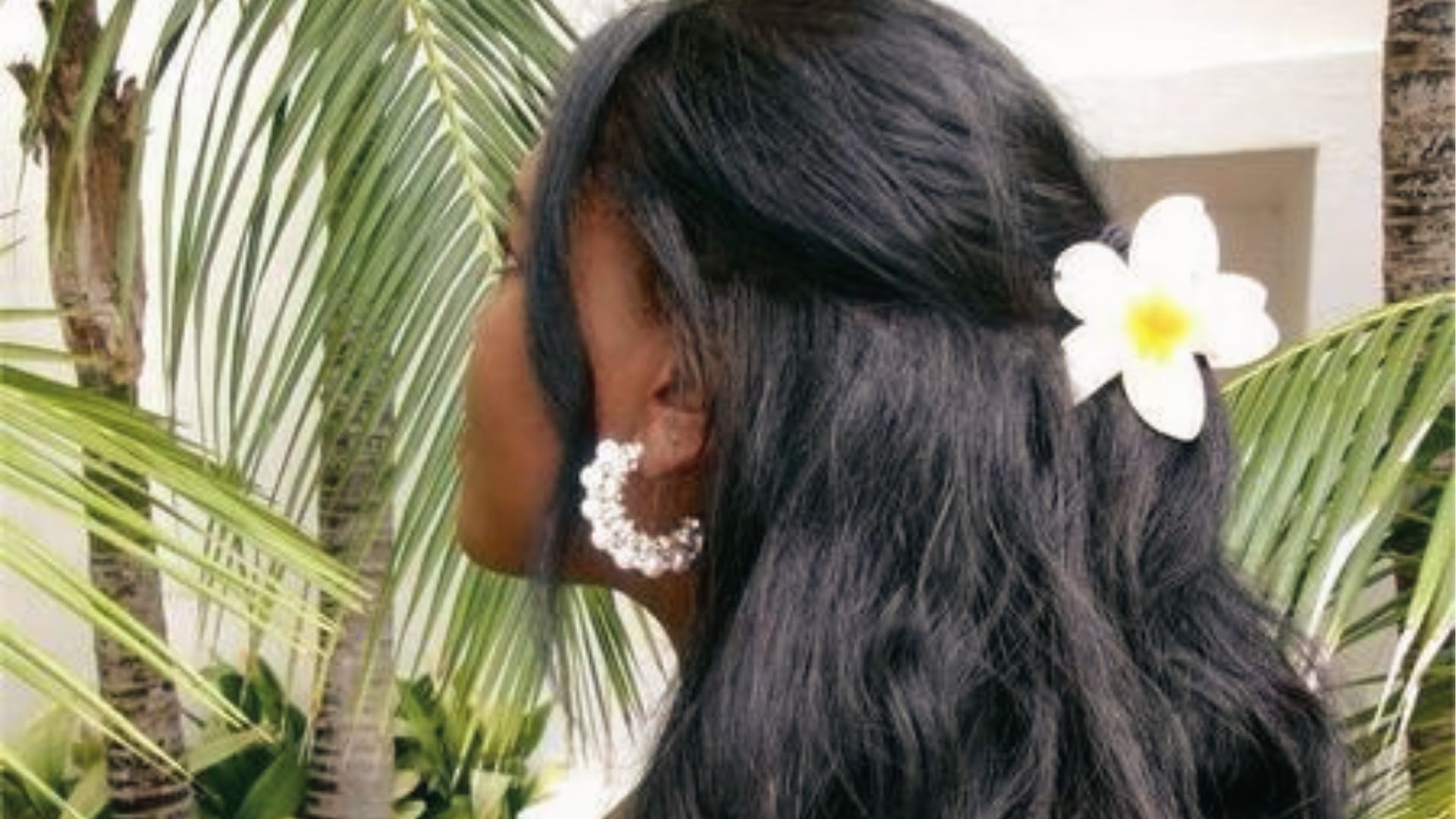 Half up half down wavy black hairstyle with white flower clip and pearl hoops in tropical setting