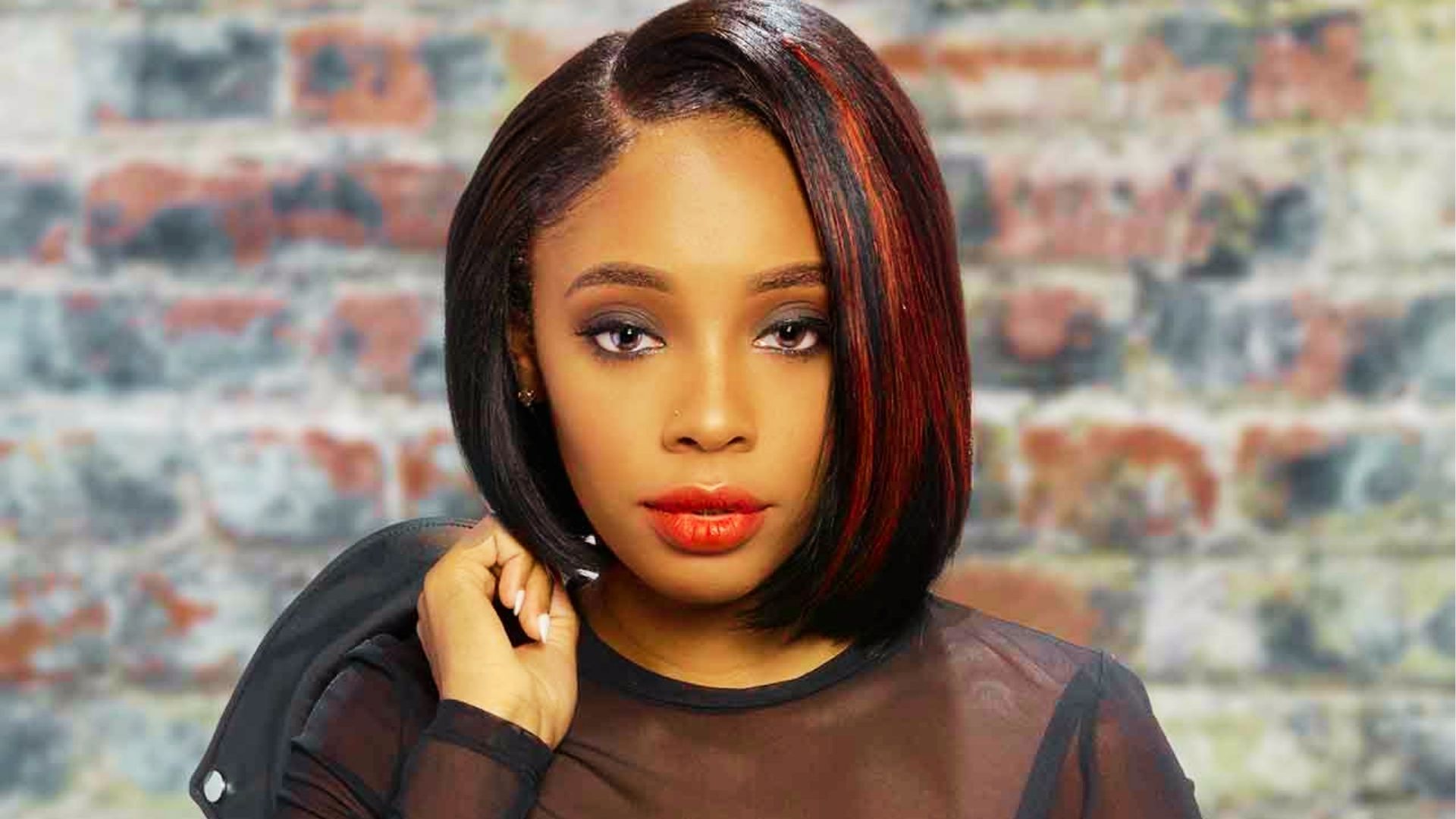 Woman with sleek bob haircut featuring red highlights wearing sheer black top against brick wall