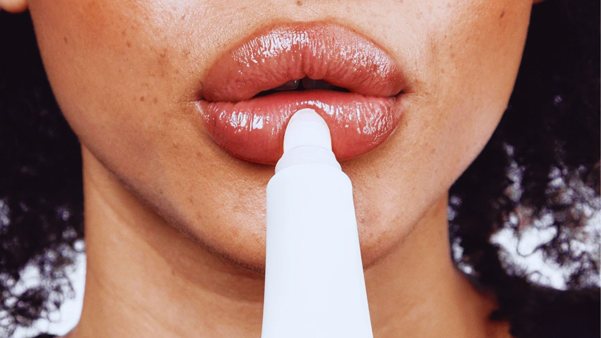 Women with curly hair apply white lip plumper tube to glossy nude lips for fuller appearance and hydration