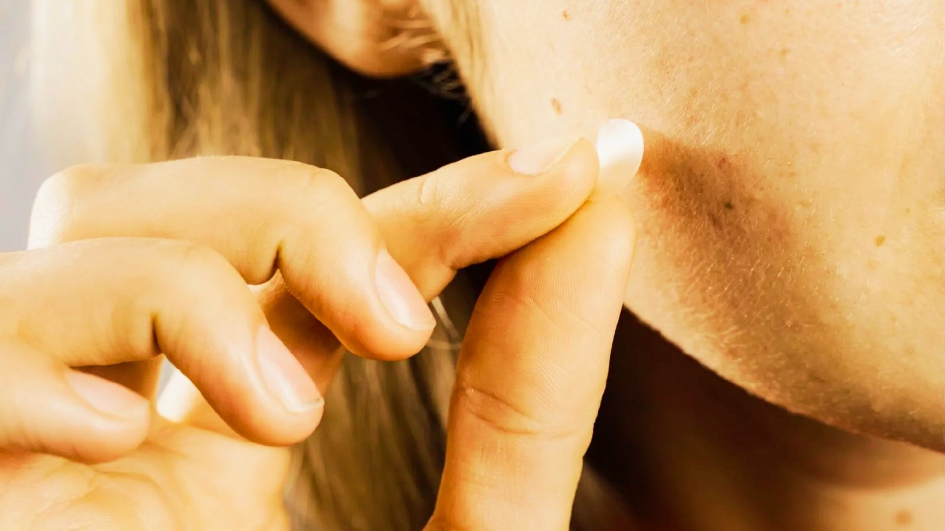 Woman applying hydrocolloid pimple patch on clean skin with bright skincare-focused bathroom lighting and mirror.
