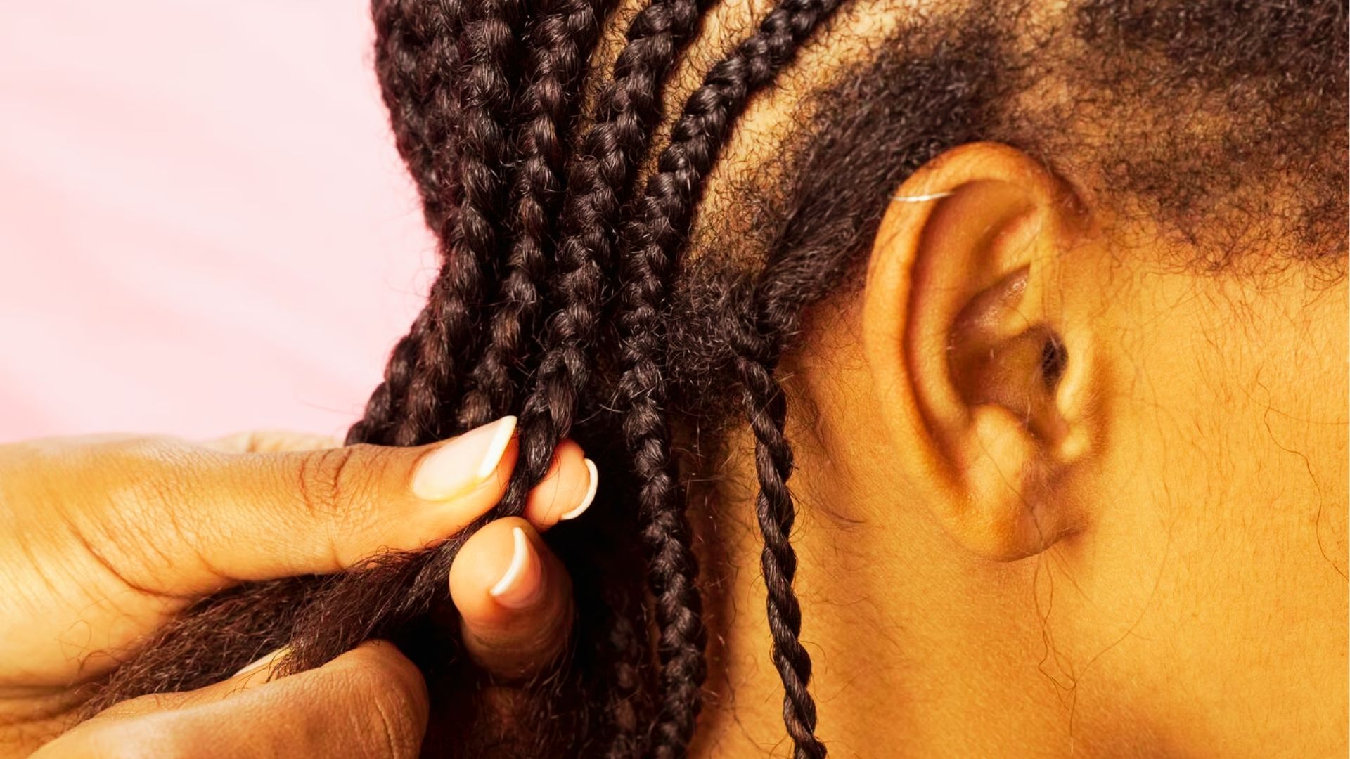 Close up of hands braiding natural hair into neat cornrows against soft pink background