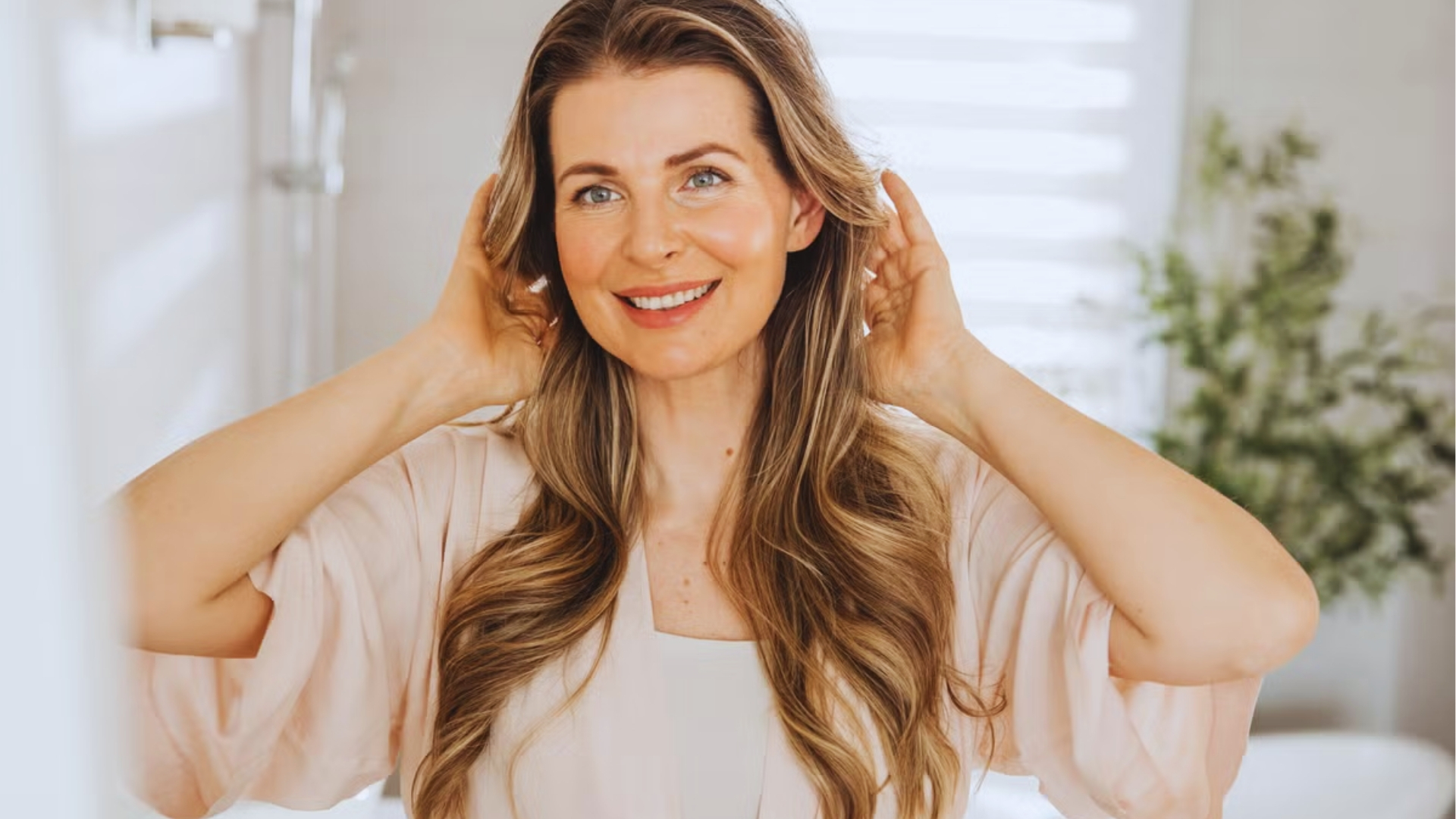 Smiling woman with wavy blonde highlights runs hands through voluminous hair, showing shine after vitamin E oil treatment.