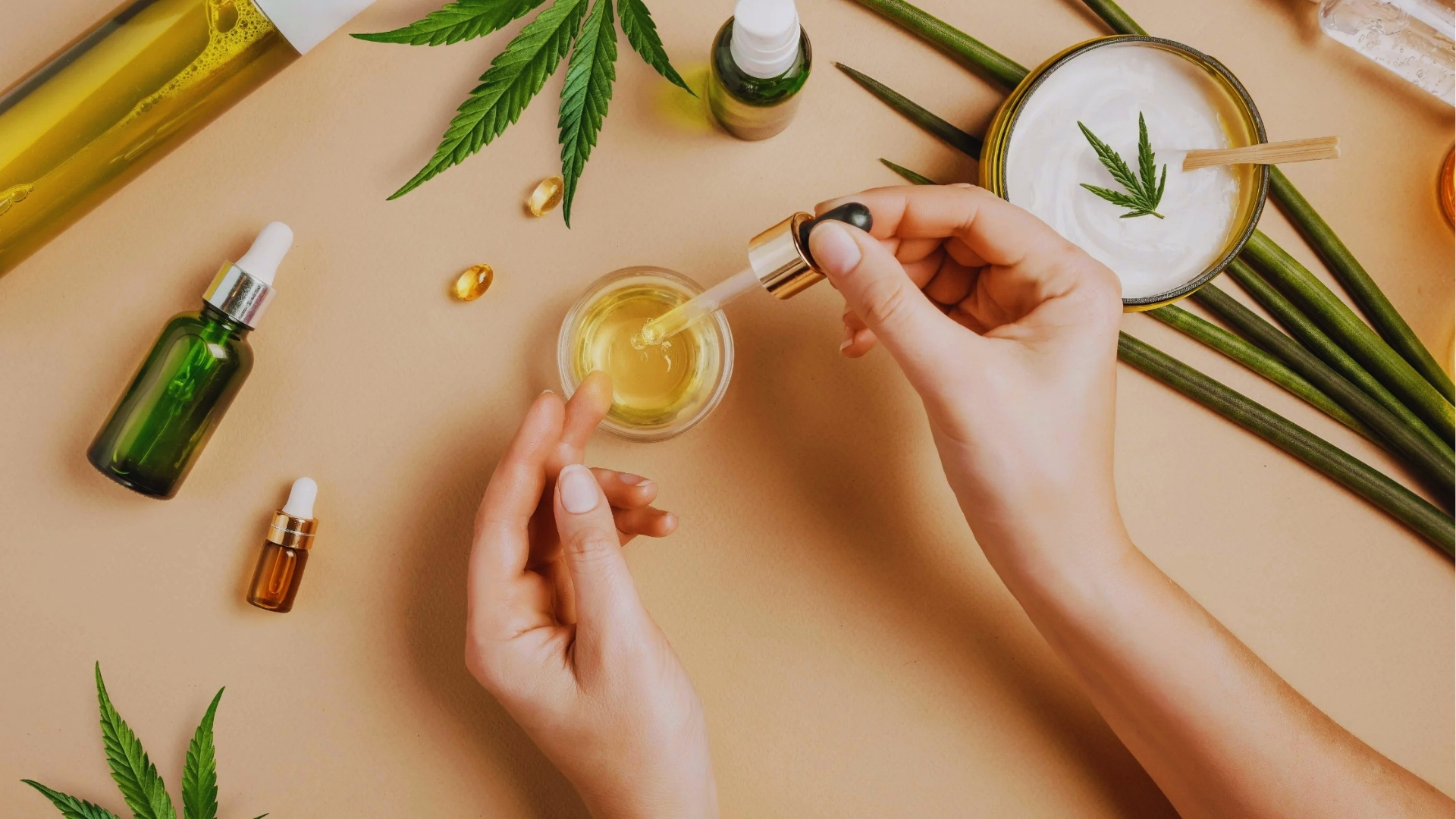Flat lay of vitamin E oil bottles, capsules, hemp leaves, and cream on beige background for natural hair care routine.