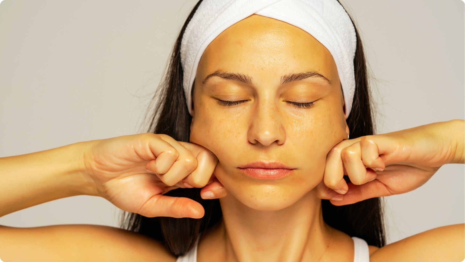 Woman with eyes closed wearing a white headband, using her knuckles to massage both cheeks.