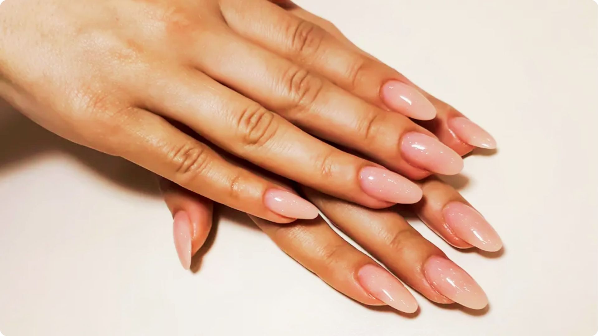 Two hands with long almond-shaped nails in a glossy nude shade.