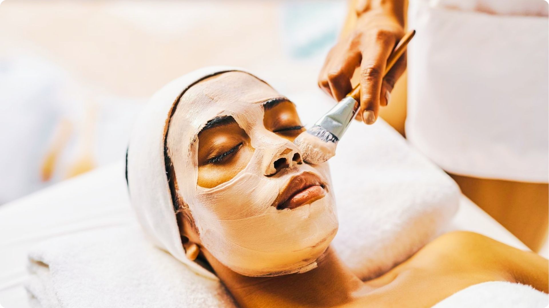 Esthetician applying a clay face mask with a brush to a client lying down with a towel wrap.