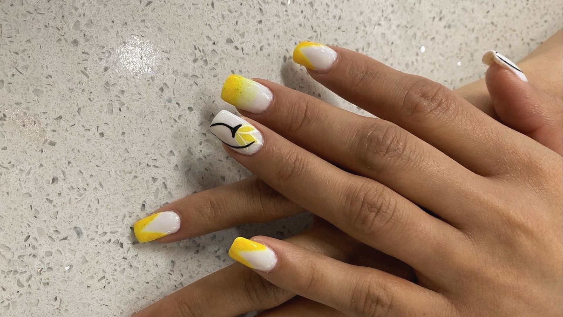 Square nails with neon yellow French tips and lemon slice art on white base, on terrazzo counter