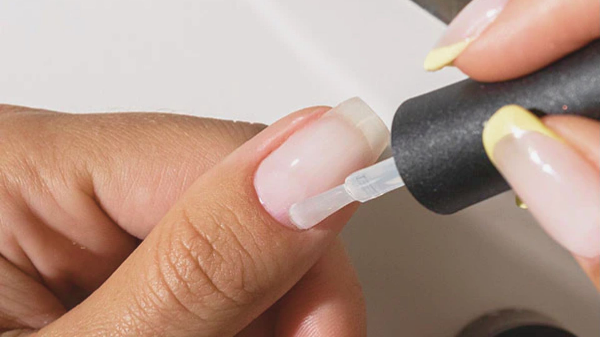 Close-up of a nail technician applying clear gel base coat to a natural fingernail using a brush.