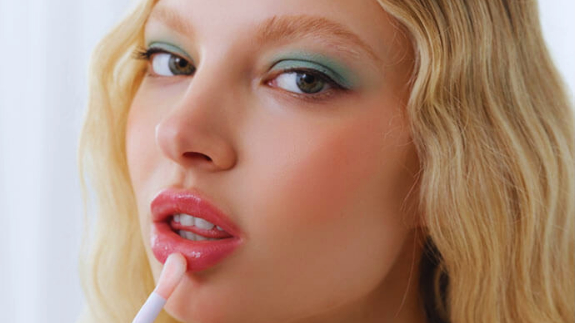 Blonde woman with green eyeshadow applies pink lip plumper gloss to lips, showing beauty routine for volume.