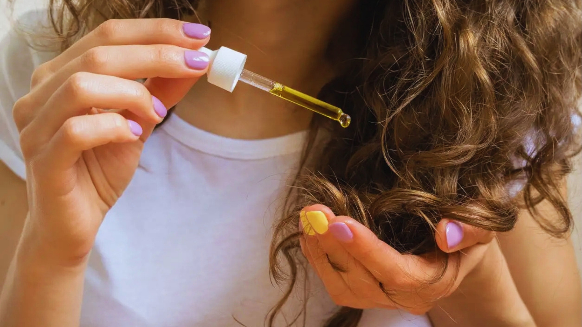 Woman with lavender and yellow nails uses dropper to apply golden vitamin E oil to curly hair for scalp nourishment