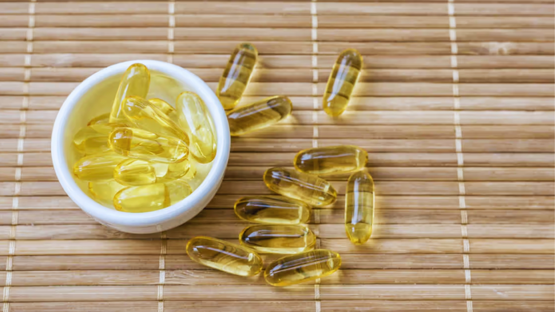 White bowl of yellow vitamin E capsules on bamboo mat, some spilled, highlighting supplement for hair growth.