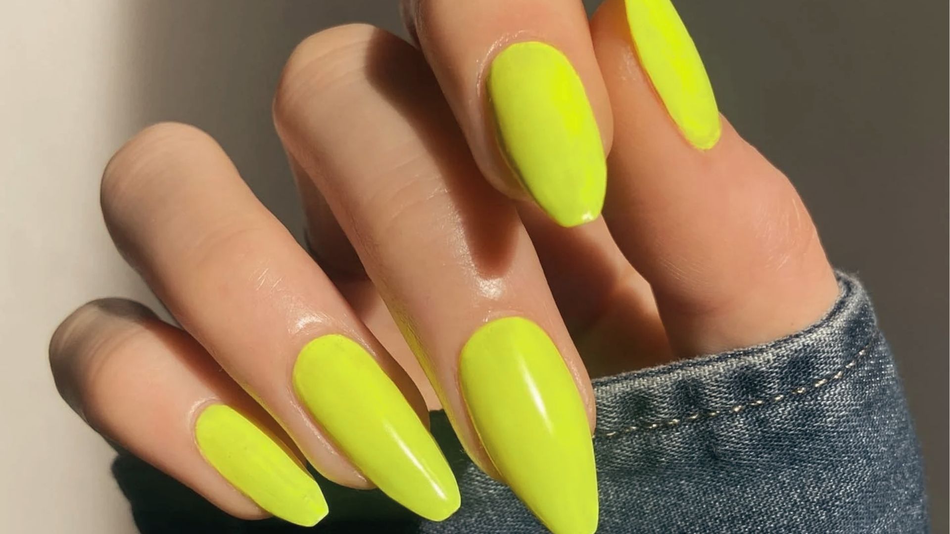 Long almond nails in bright neon yellow, sunlight on hand with denim sleeve, glossy finish