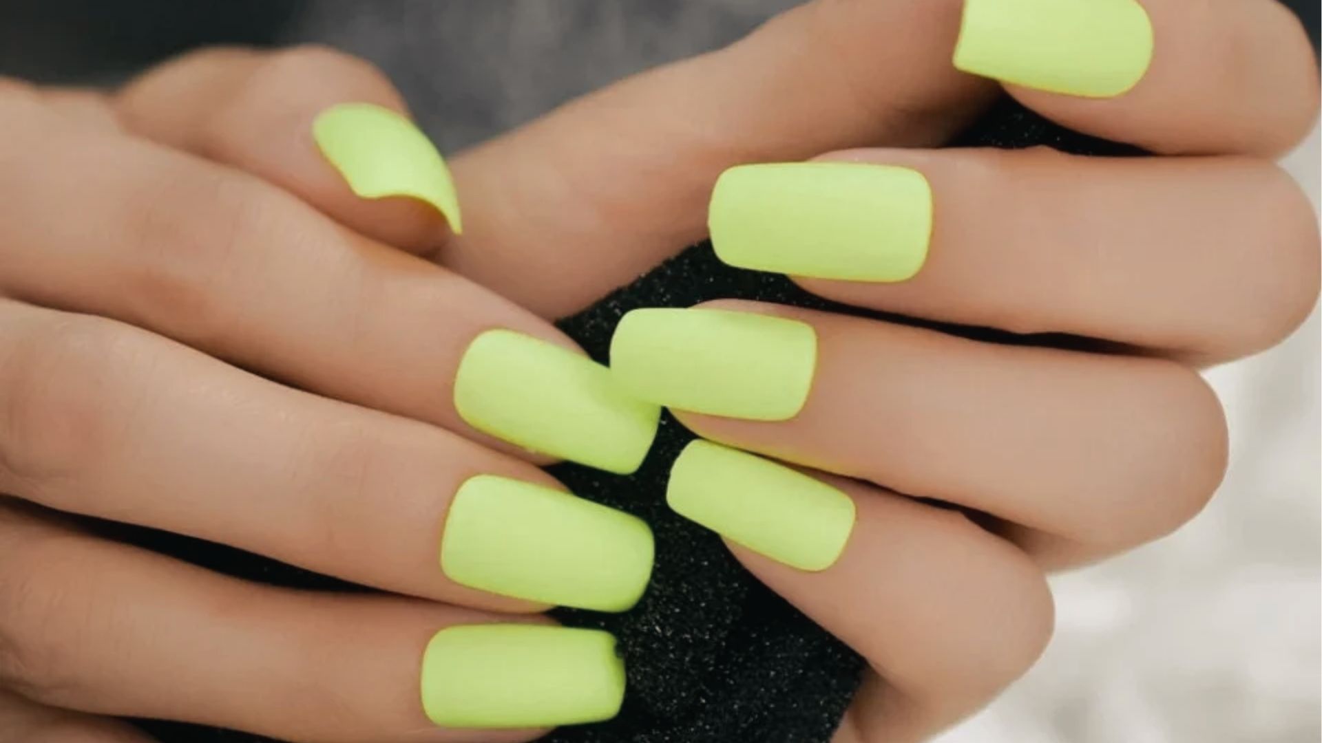 Short square nails in matte neon yellow, hands crossed holding black sponge, smooth finish