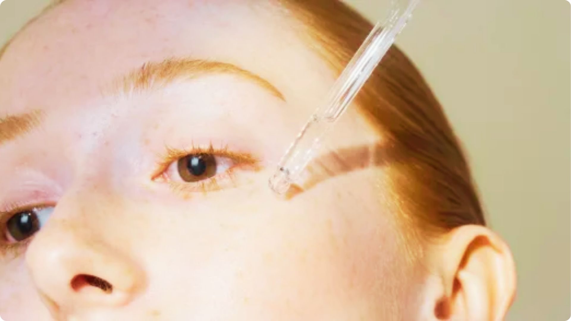 Close-up of a dropper applying Retinoids serum to a person’s forehead near the brow.