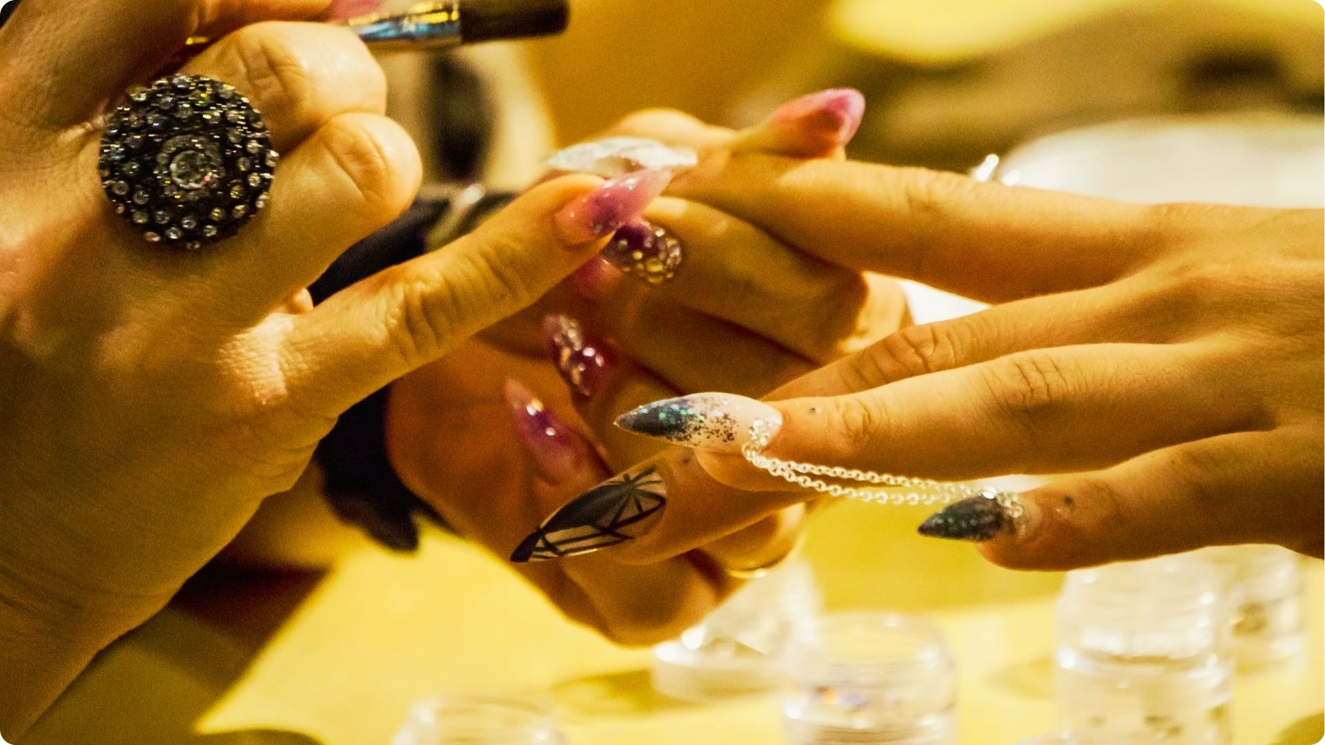 Close-up of a technician attaching a delicate chain to long, decorated stiletto nails with glitter and patterns.