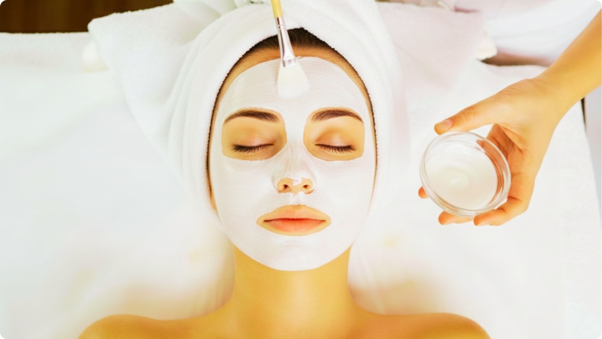 Woman receiving a white cream face mask applied with a brush while relaxing at a spa.