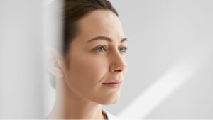A woman with clear skin stands by a window, eyes closed, embodying serenity and a frown line-free complexion.