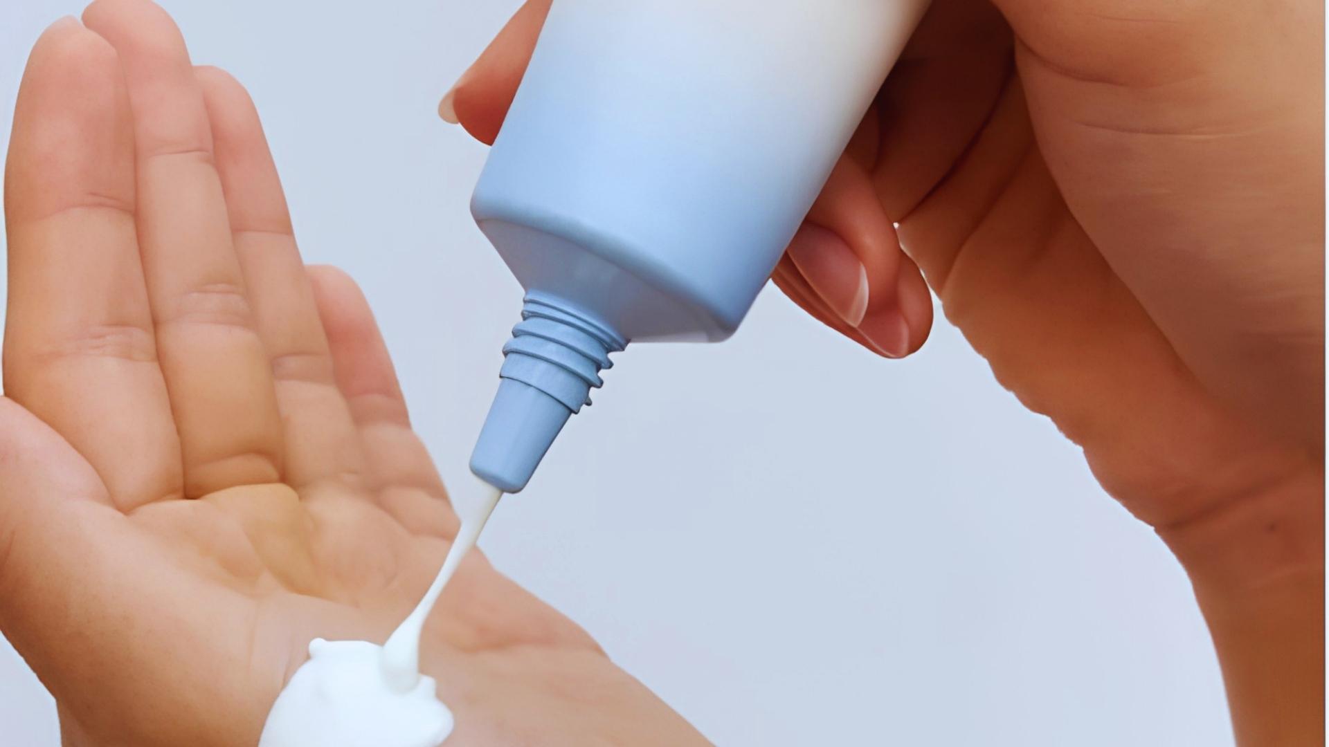Hands squeeze light blue sunscreen tube, dispensing thick white cream onto open palm against plain light blue background showing texture.