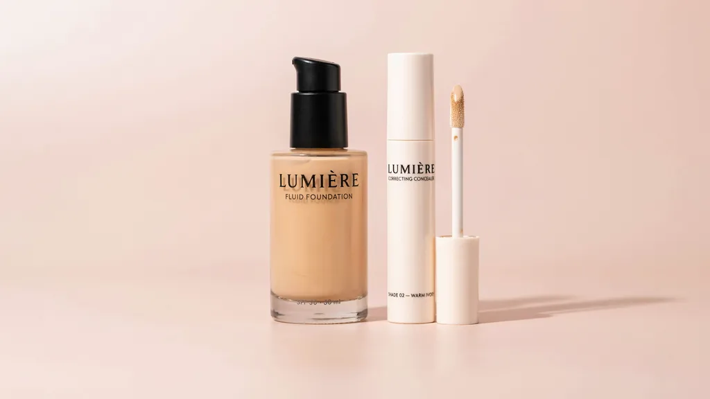 Lumière Fluid Foundation with SPF 30 in a glass pump bottle beside an open Lumière Correcting Concealer in Shade 02 Warm Ivory on a blush pink surface.